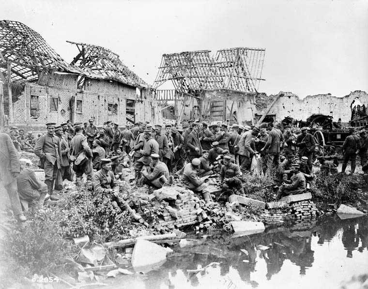 Photographie en noir et blanc – De nombreux prisonniers de guerre allemands sont rassemblés en bordure d’une rivière. Les décombres et des arbustes jonchent le sol. Les hommes sont entourés de ruines.